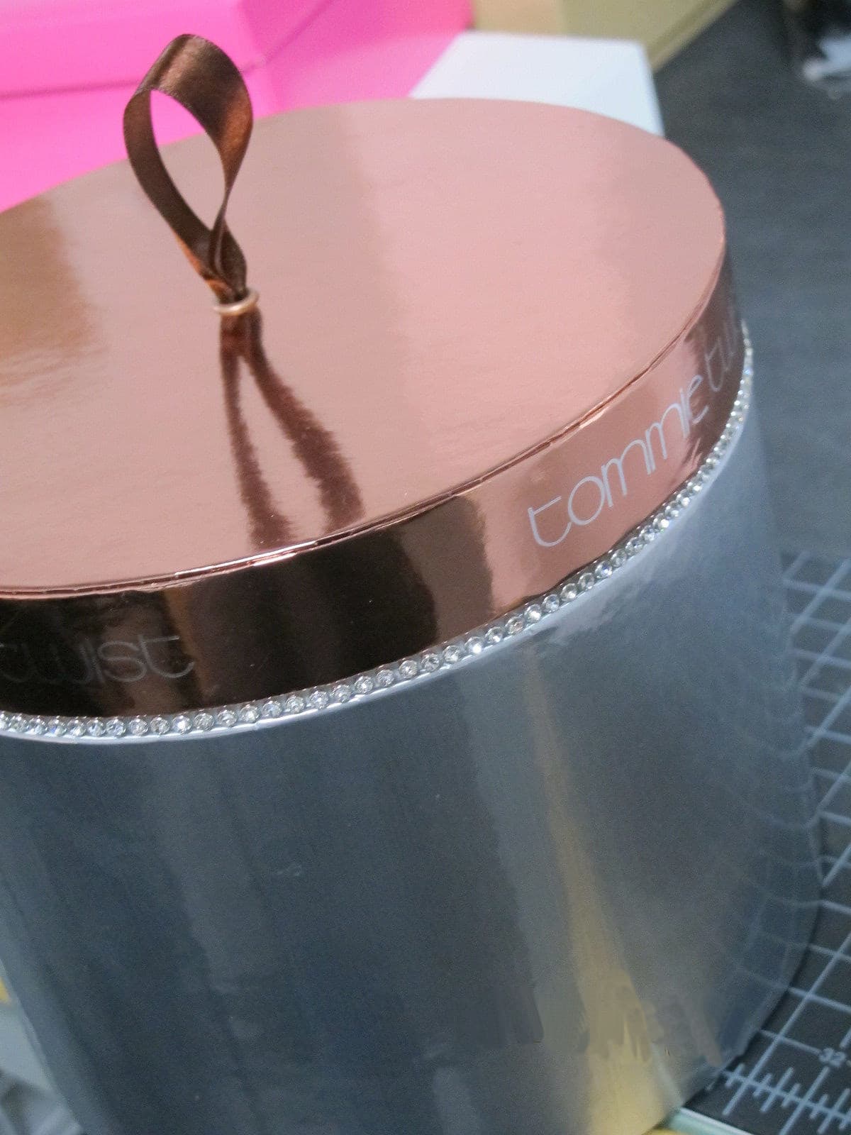 Cylindrical grey container with a reflective rose gold lid, brown ribbon loop, and rhinestone trim.